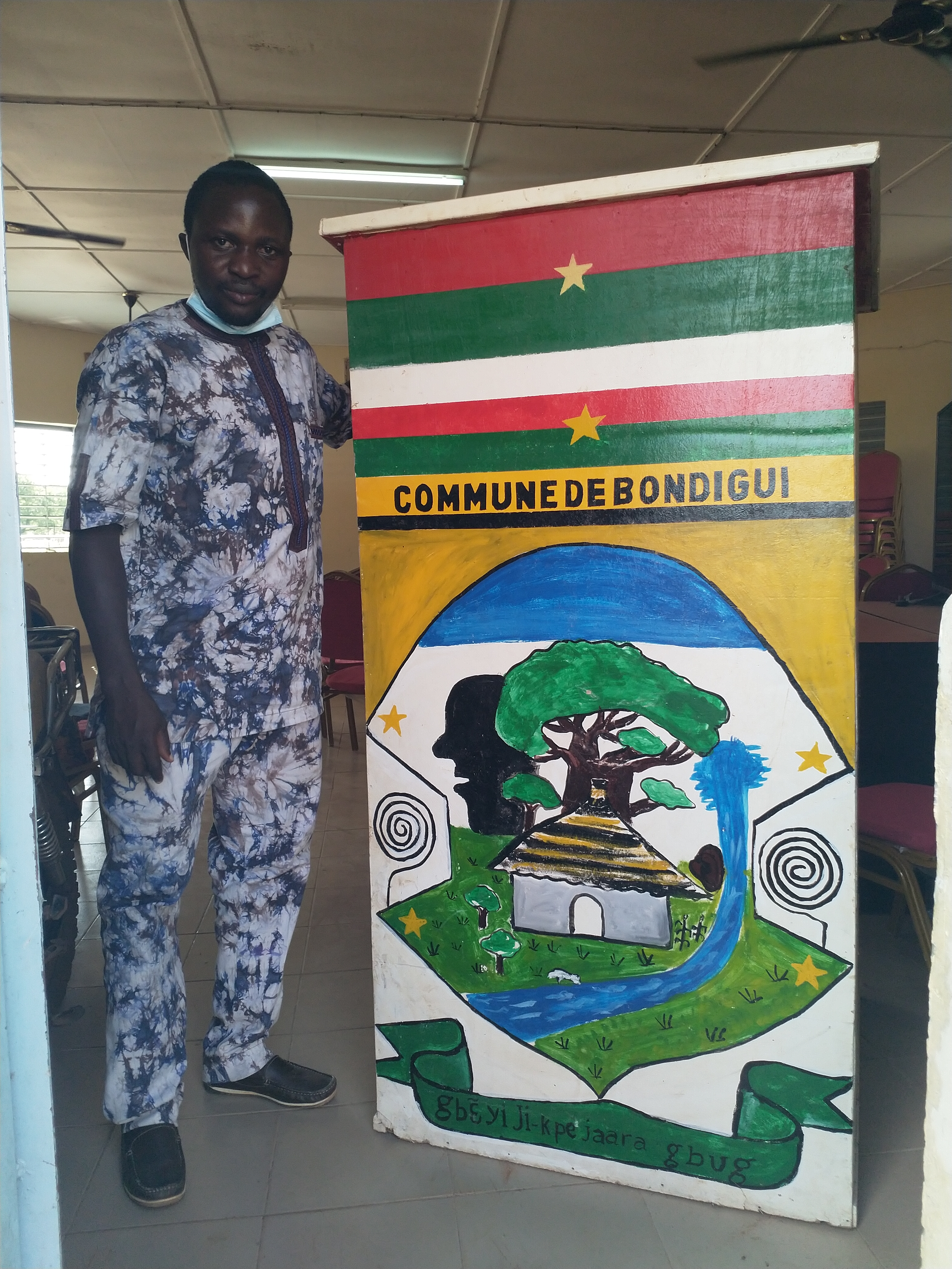 A man standing next to the coat of arms of a the municipality of Bondigui, Burkina Faso.