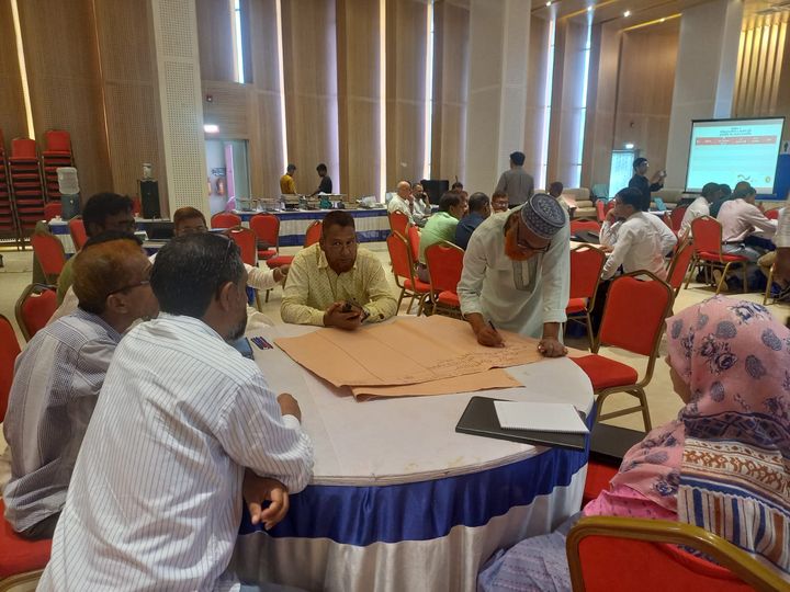 A room full of people sitting around round table, the focus is on one table where a group of men is apparently discussing things and noting them on brown big paper sheets.