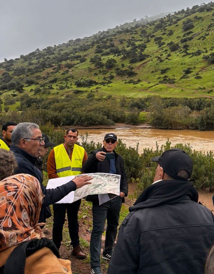 A group of people are standing around a man with a map in his hands. He is explaining something while they are all standing next to a river, on the other side of the river a hill rises.