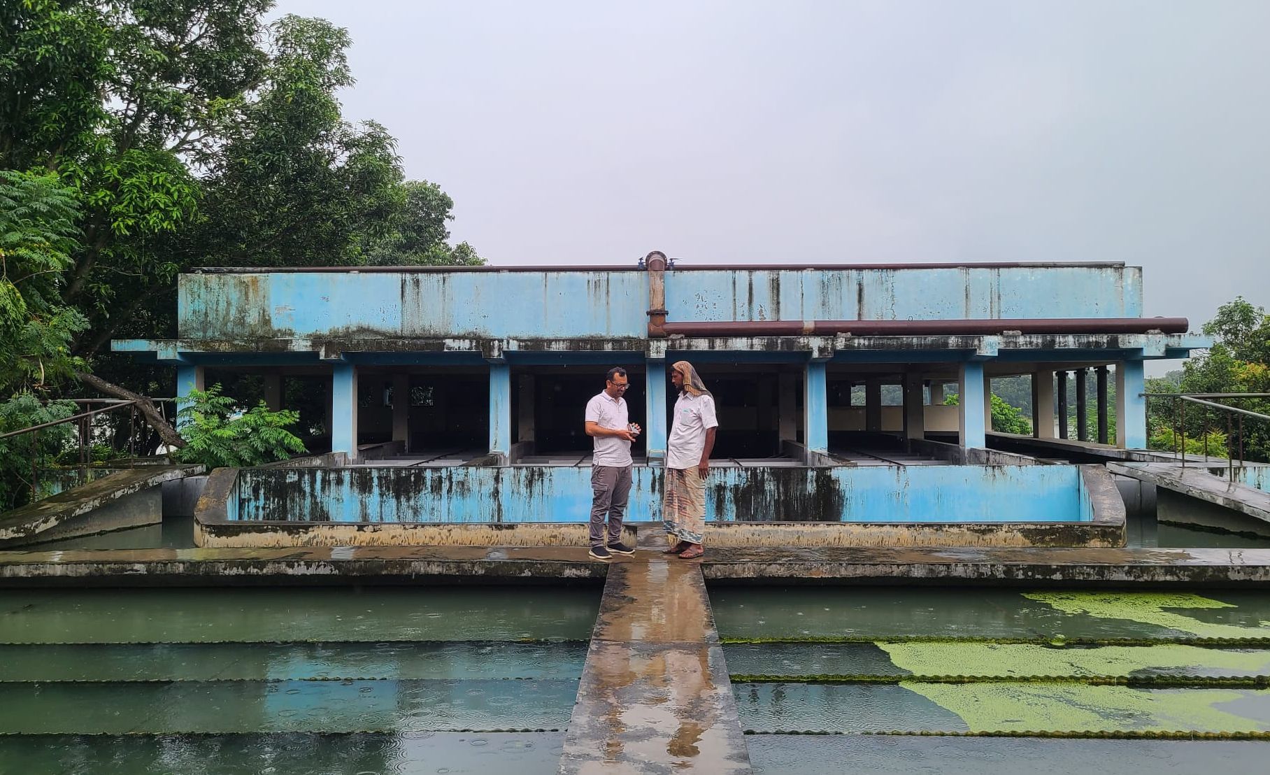 Two men facing each other, standing at in front of a blue building in the back of the picture, in front of them a well structured water basin.