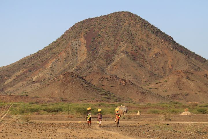 Three people balancing jerrycans on their heads walking on an arid landscape towards a brown mountain.