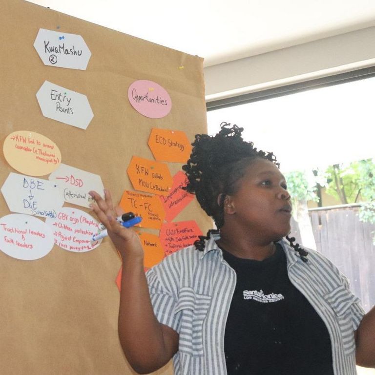 A black woman is standing in front of a chart with written cards pinned to it, facilitating a workshop.