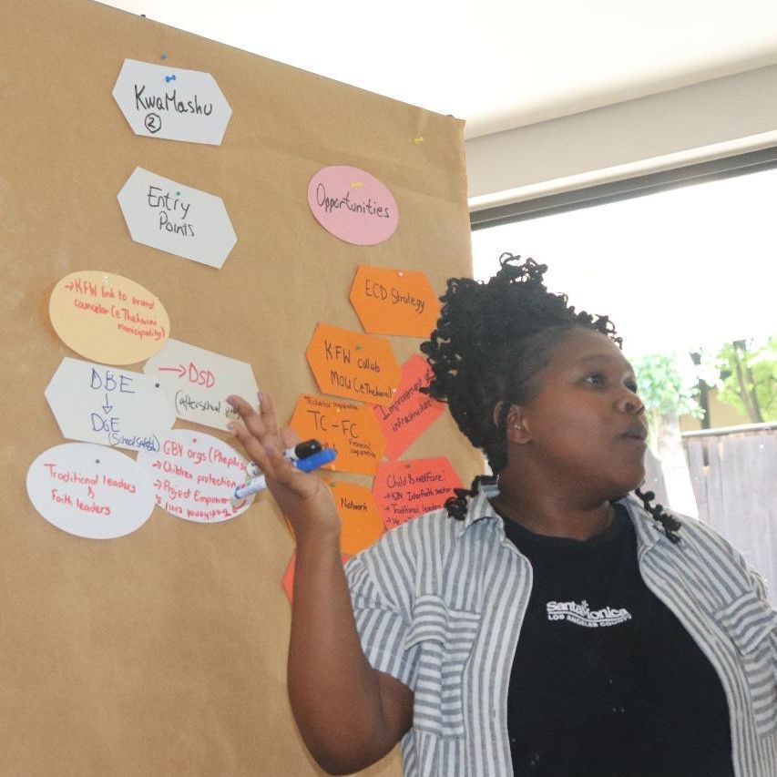 A black woman is standing in front of a chart with written cards pinned to it, facilitating a workshop.