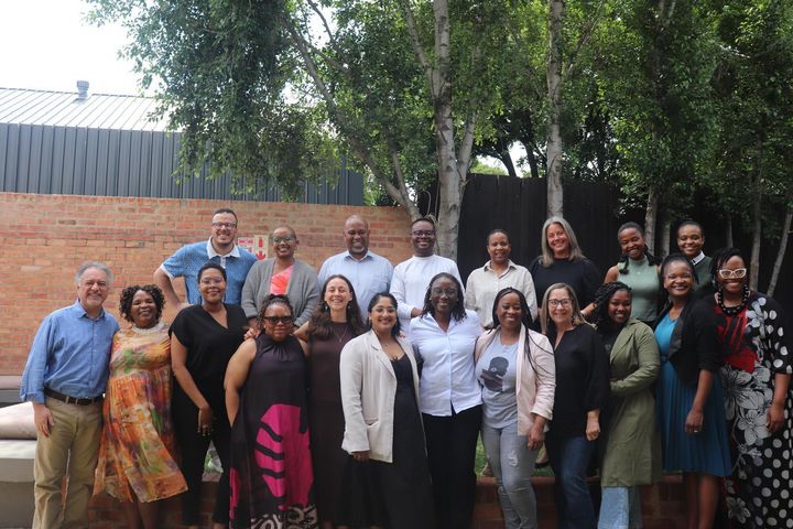 A group of about smiling 20 people posing for a picture under a big tree with a wall in their back.