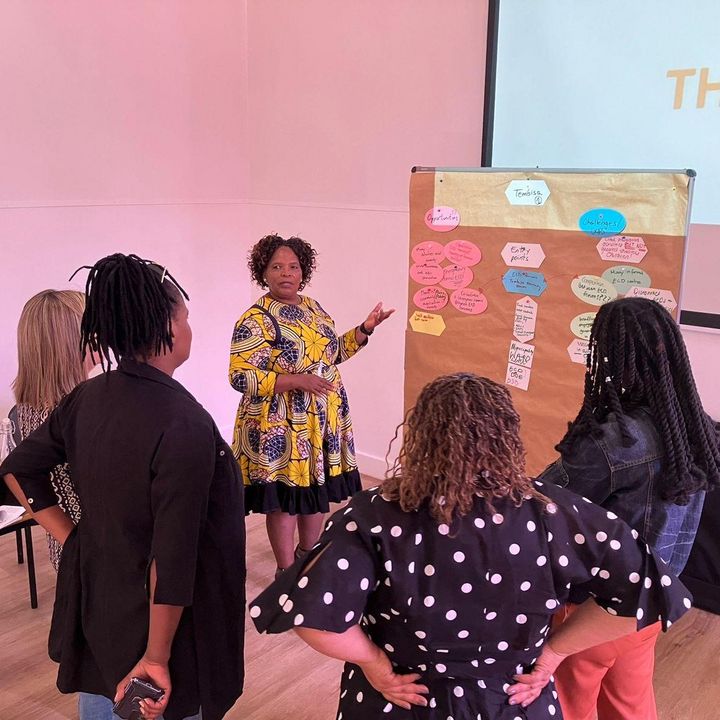 A group of mostly black women standing in front of a chart, a black female facilitator is pointing on the chart and talking to the other women.