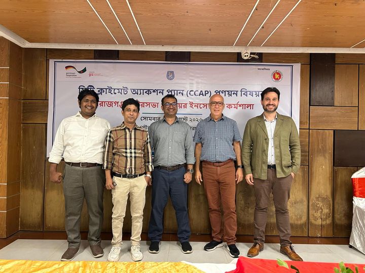A group of five men in casual business clothes standing in front of a banner with logos and a text in bangla.