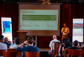 A man with a microphone in his hand standing in front of a screen between two banners, in front of many people seated in chairs.
