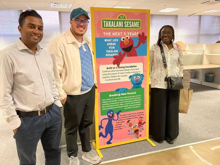 Two men and a woman, smiling, standing around a roll up banner informing about the South African Takalani Sesame (Sesame Street) and how it will start addressing topics like bias and prejudice.