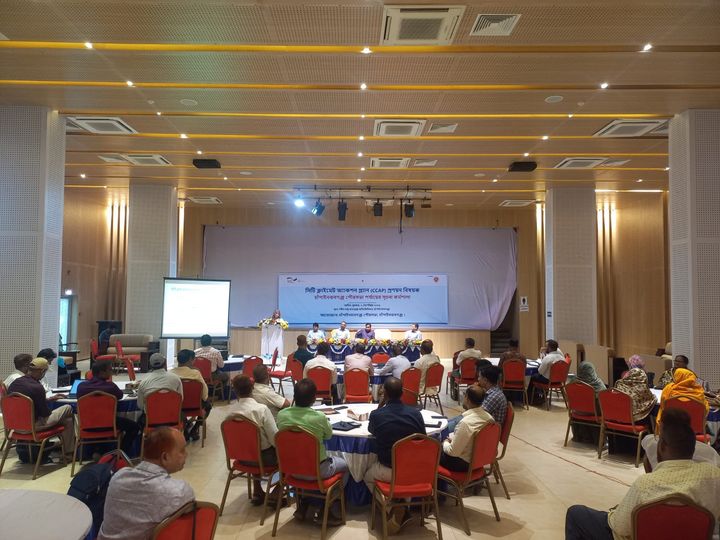 A big conference room with a panel of speakers in the back and many people sitting around different round tables paying attention to the panel.