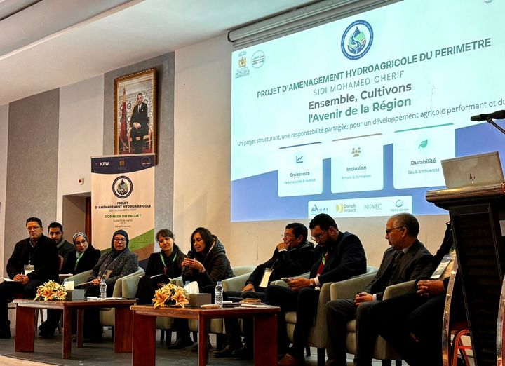 A group of people sitting on a podium in a formal setting, tables in front of them and a presentation projected to the wall at their back.