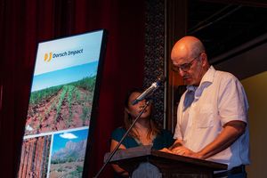 A man standin at a podest looking down to his script, a woman in the back next to a banner.