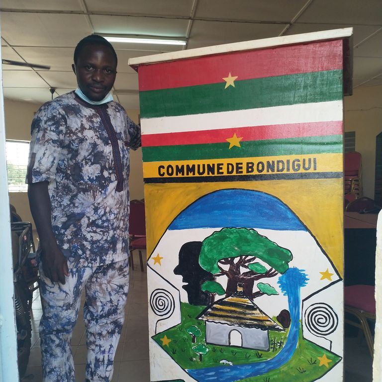 A man standing next to the coat of arms of a the municipality of Bondigui, Burkina Faso.