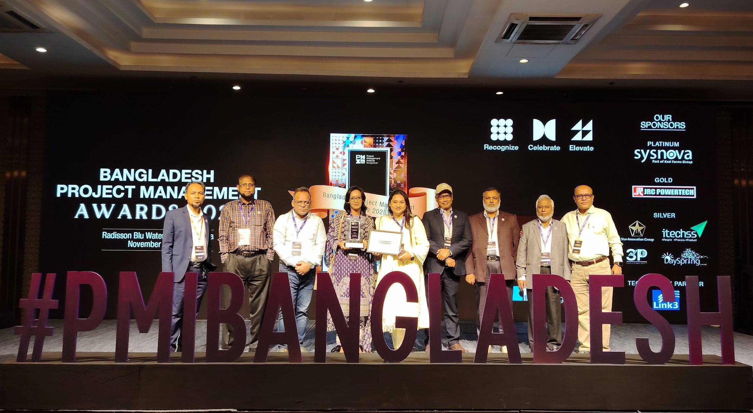 The delegation of the CReLIC team with the PM Award and diploma behind a big board saying #pmibangladesh 