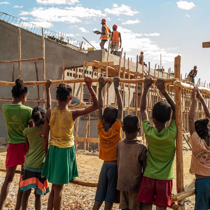 A group of children seen from the back, observing the construction of their new school behind a fence.