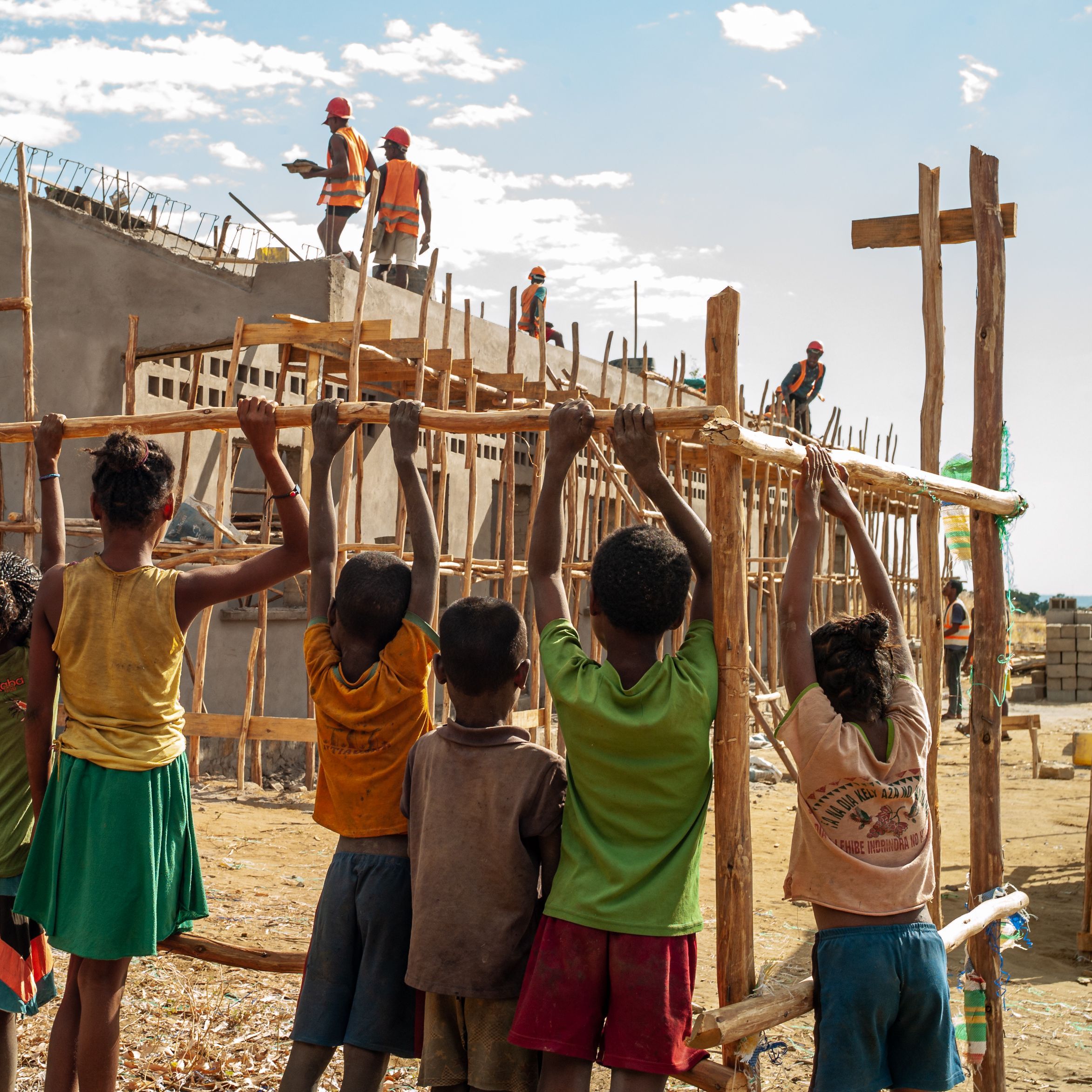A group of children seen from the back, observing the construction of their new school behind a fence.